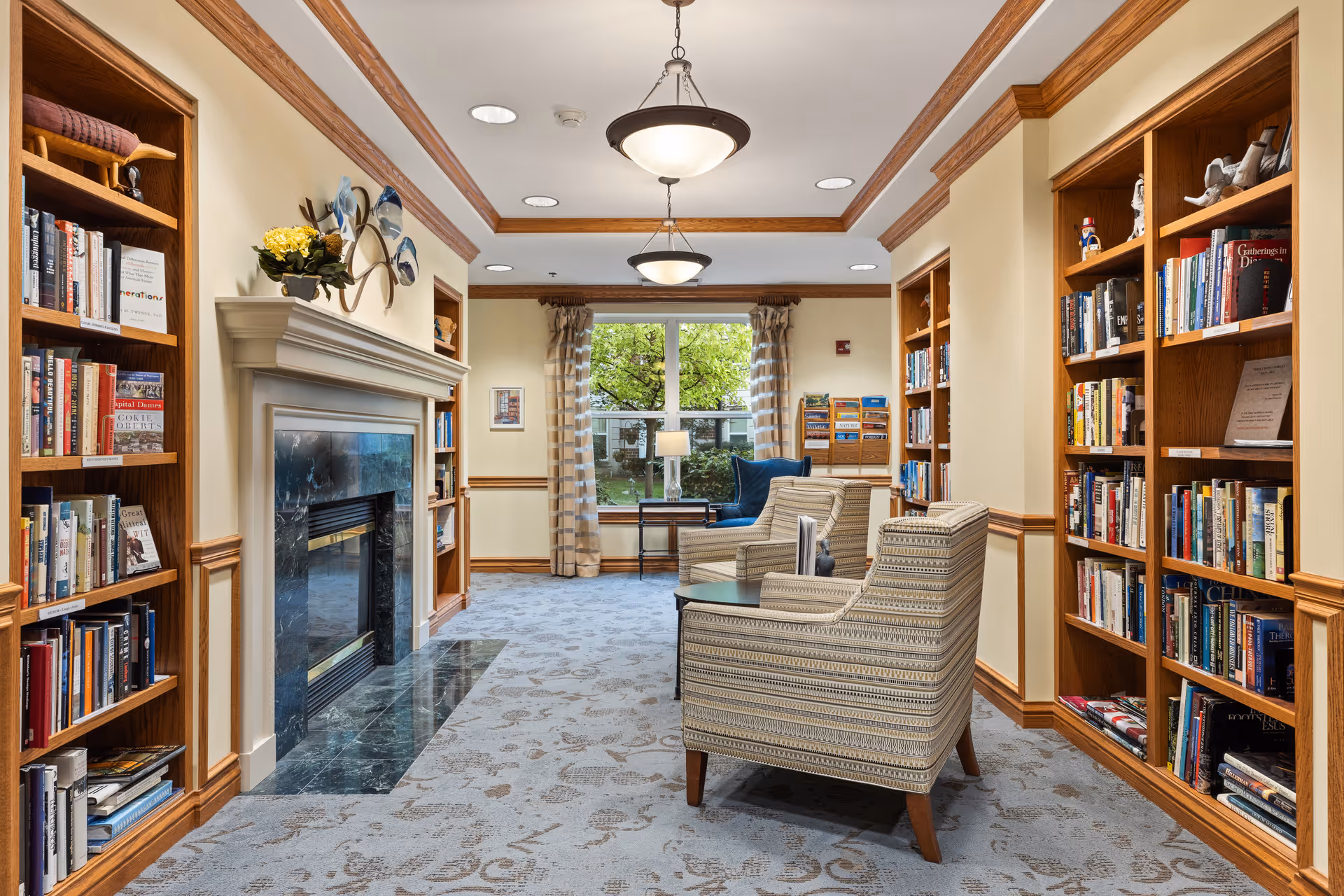 Cozy library-style common room with built-in bookshelves, a fireplace, and upholstered armchairs facing a window.