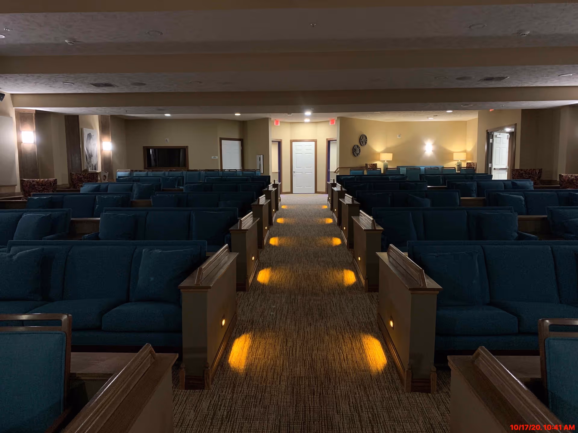 Interior view of a large room with multiple rows of blue cushioned pew-style seating arranged on either side of a central carpeted aisle with small lights along the sides. The room has beige walls, wall sconces, and lamps providing warm lighting. There are doors and decorative wall clocks visible at the far end of the room.