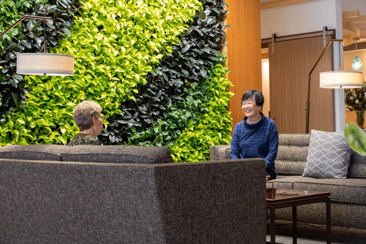 Two elderly women sitting and talking in a modern lounge area with gray sofas, a wooden coffee table, and a vibrant green vertical plant wall behind them. The room is softly lit with floor lamps and has a warm, welcoming atmosphere.