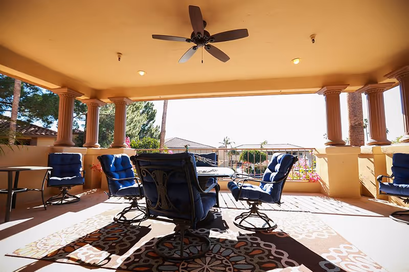 Covered outdoor patio area with cushioned blue swivel chairs arranged around a rectangular table, ceiling fan above, and decorative columns supporting the roof. The patio overlooks a landscaped area with trees and rooftops in the background.