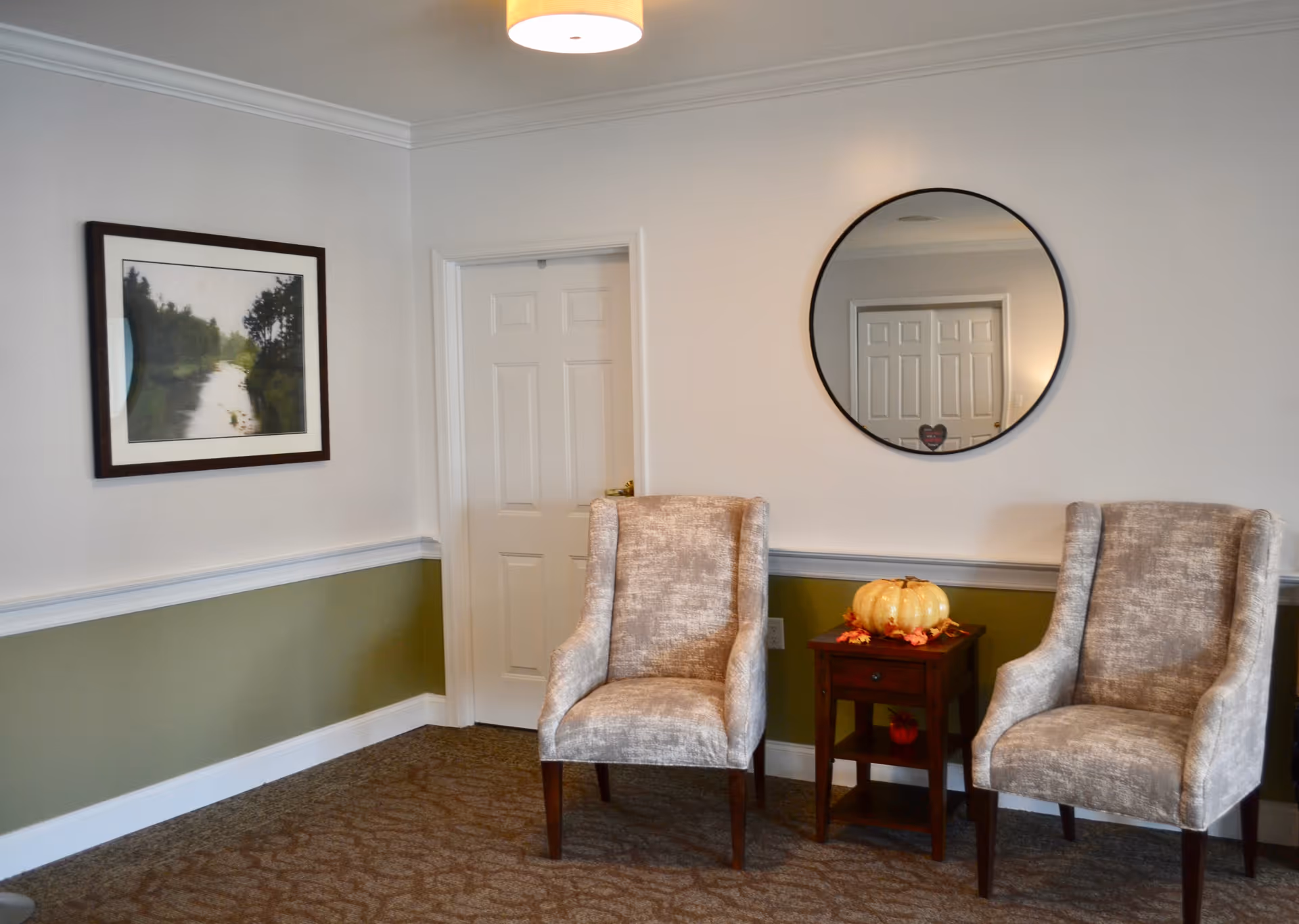 A cozy seating area with two beige upholstered armchairs separated by a small wooden side table decorated with a pumpkin and autumn leaves. A round mirror hangs on the wall above the table, reflecting a closed white door. To the left, there is a framed landscape painting on the wall. The lower half of the wall is painted green with white trim, and the upper half is light-colored.