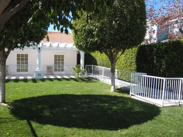 A well-maintained outdoor garden area with green grass, two trees casting shadows, a white metal railing ramp, and a white building with large windows and a red-tiled roof in the background.