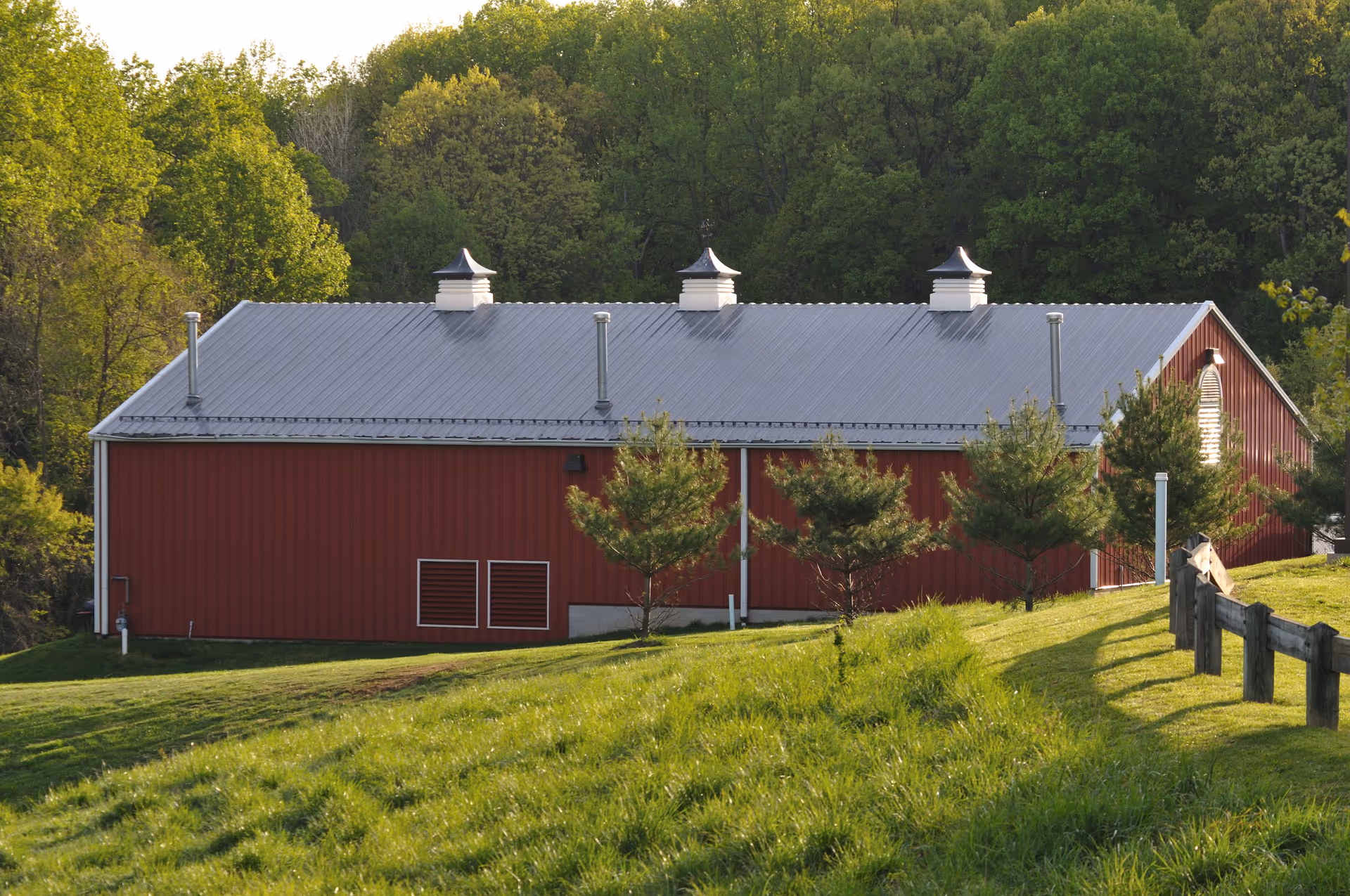 A red barn-style building with a silver metal roof and three cupolas, surrounded by green grass and small trees, with a wooded area in the background.