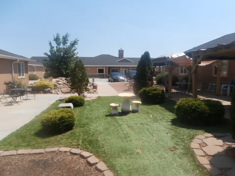 Outdoor courtyard area of an assisted living facility with green grass, small bushes, a tree, and a stone table with three mushroom-shaped stools. There are buildings surrounding the courtyard with windows and a covered patio area with chairs. A car is parked near the entrance of the building in the background under a clear blue sky.