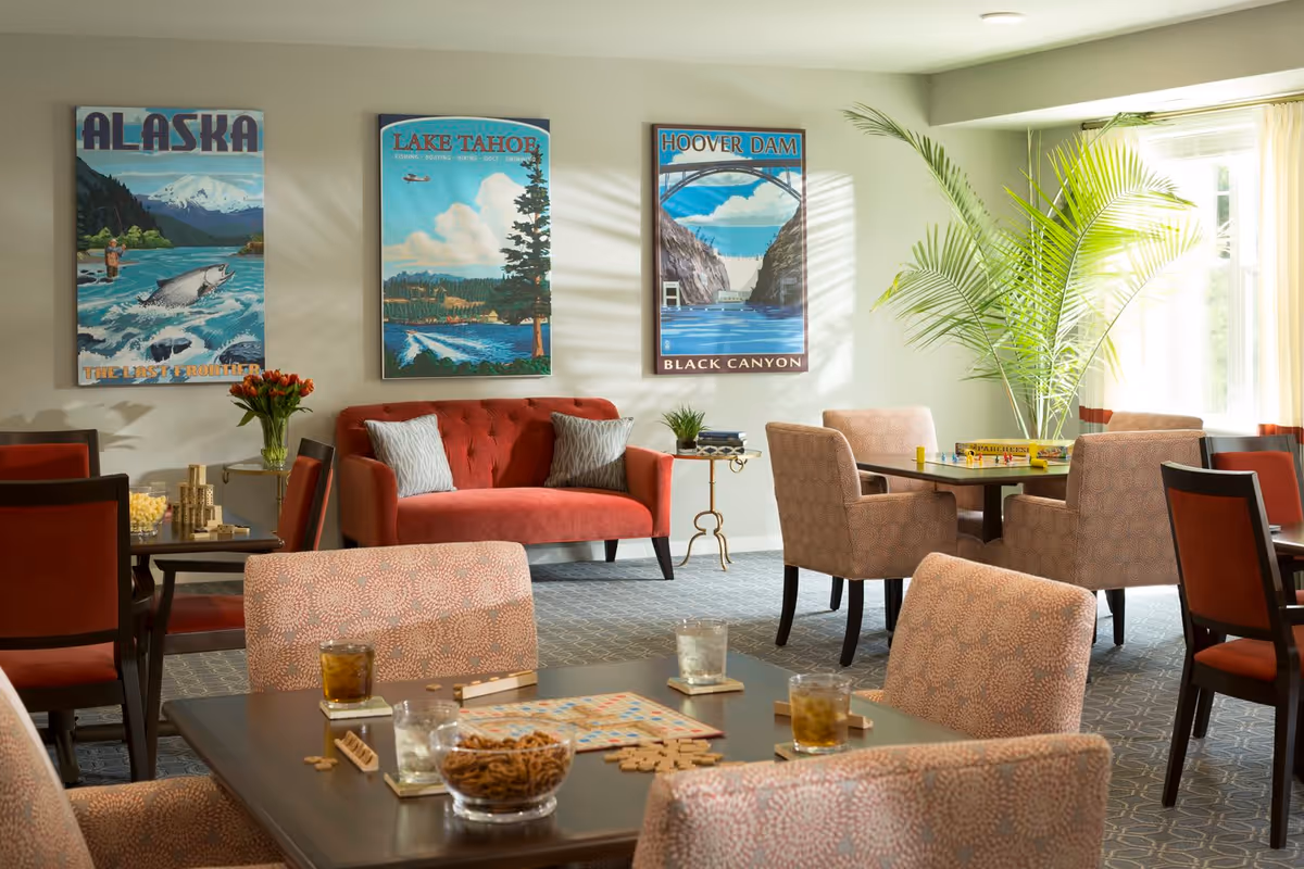 A bright and cozy common area in a senior living facility featuring several tables and chairs arranged for social activities. One table has a Scrabble game set up with drinks and snacks, while another table has a board game. The room includes a red loveseat with two pillows, a side table with a plant, and three colorful travel posters on the wall depicting Alaska, Lake Tahoe, and Hoover Dam. A large window with sheer curtains allows natural light to fill the space, and a tall green plant adds a touch of nature.