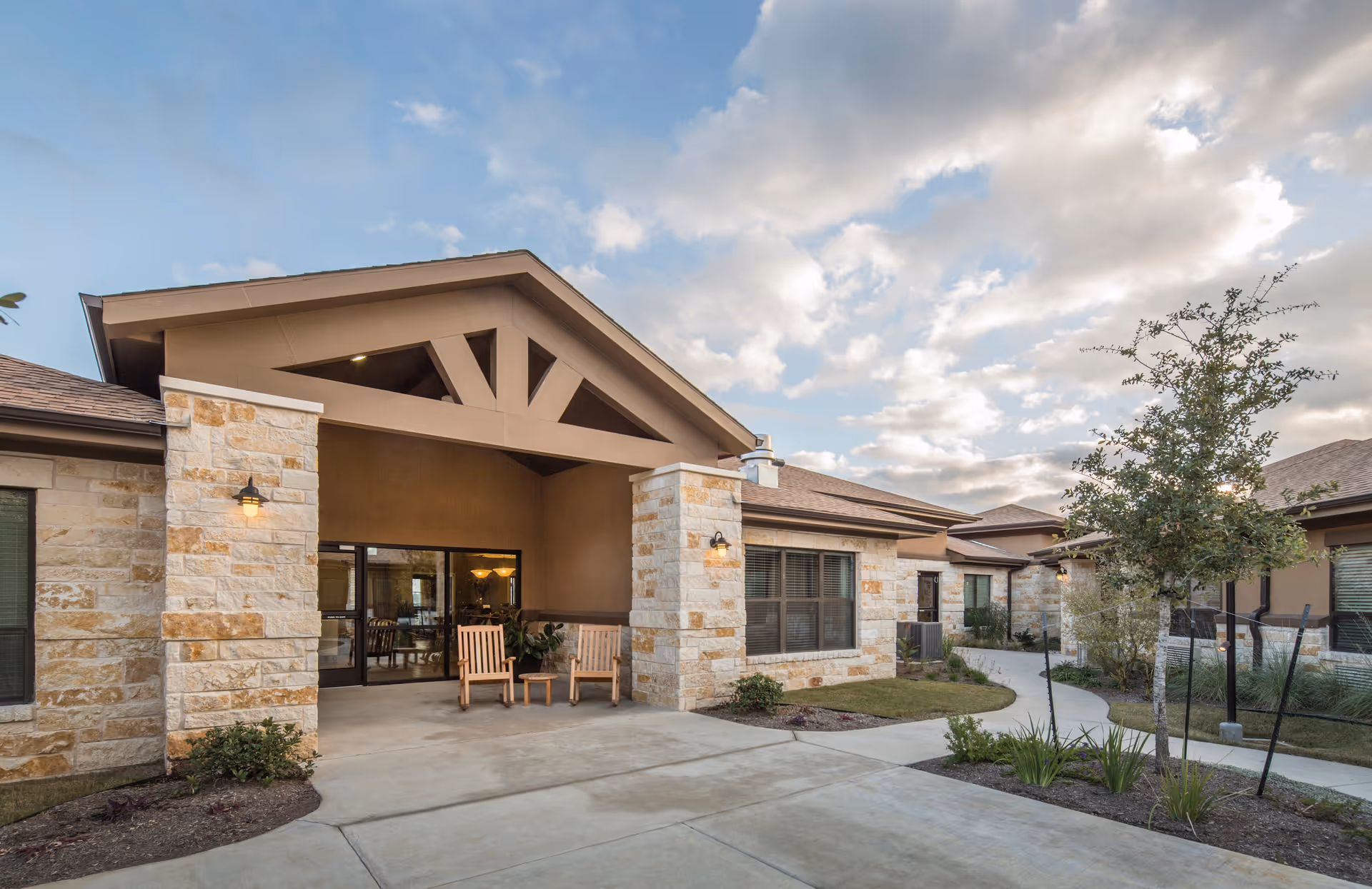 Exterior view of Park Manor Bee Cave facility showing a stone and stucco building with a covered entrance, two wooden chairs and a small table on the porch, landscaped garden beds, a curved concrete walkway, and a partly cloudy sky.