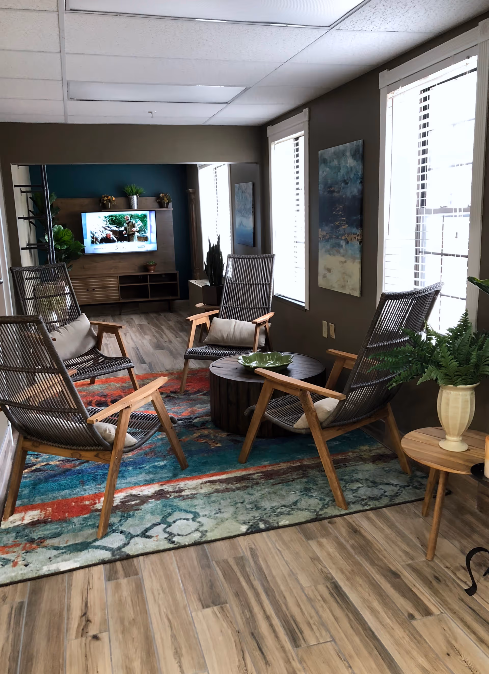 A cozy seating area with four wooden and wicker chairs arranged around a round wooden coffee table on a colorful patterned rug. The room has wood-look flooring, two large windows with white blinds, a small side table with a potted fern, and a wall-mounted TV in the background with plants on the TV stand.