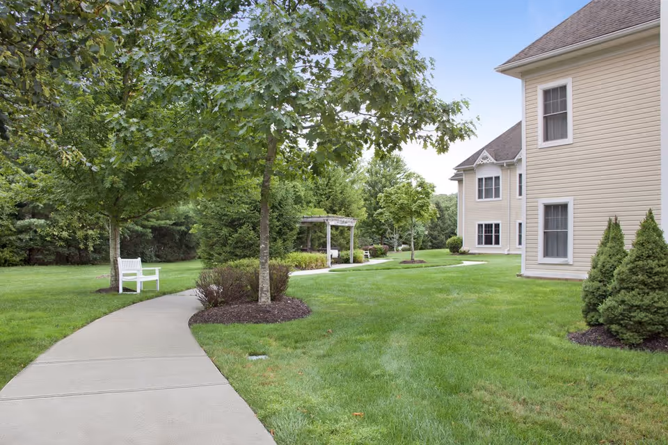 A paved walkway curves through a well-maintained grassy area with trees and shrubs. There is a white bench along the path and a wooden pergola further down the walkway. On the right side, there is a beige building with multiple windows. The sky is clear and blue.