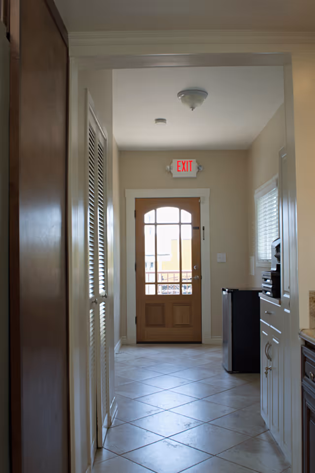 A narrow hallway with tiled floor leading to a wooden door with glass panels and an illuminated red EXIT sign above it. On the right side, there is a small black refrigerator and white cabinets with a window above them. On the left side, there are white louvered closet doors.