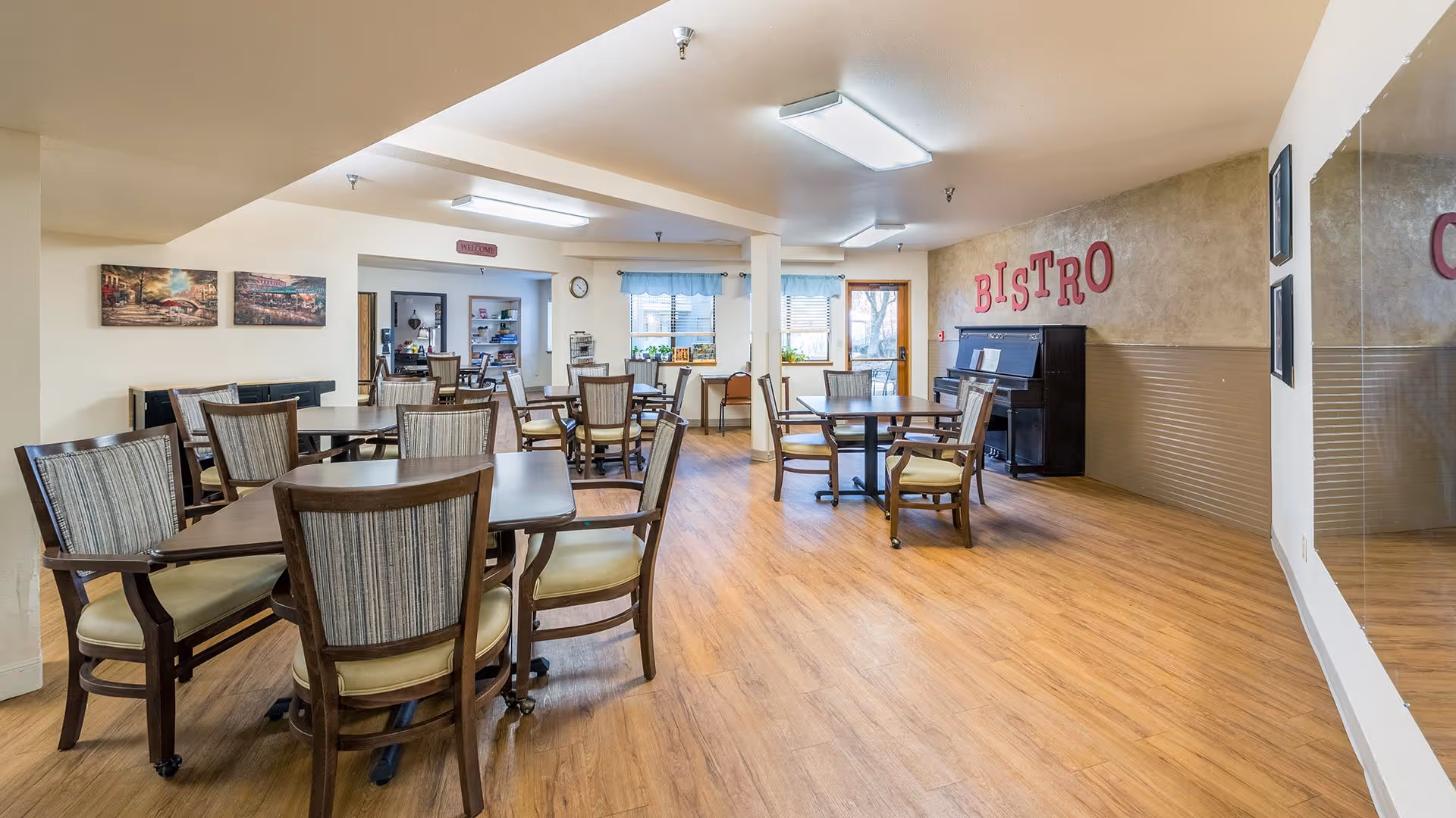 Spacious dining room with multiple tables and chairs, a piano under a 'BISTRO' sign, and wood flooring.