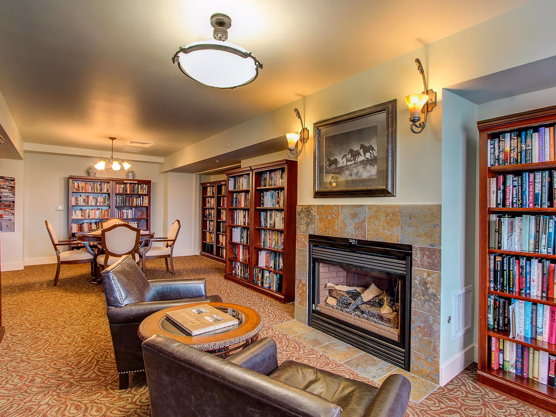 A cozy library room with bookshelves filled with books lining the walls, a fireplace with a framed picture of horses above it, two leather armchairs facing a round wooden table with books on it, and a dining table with chairs in the background under warm lighting.