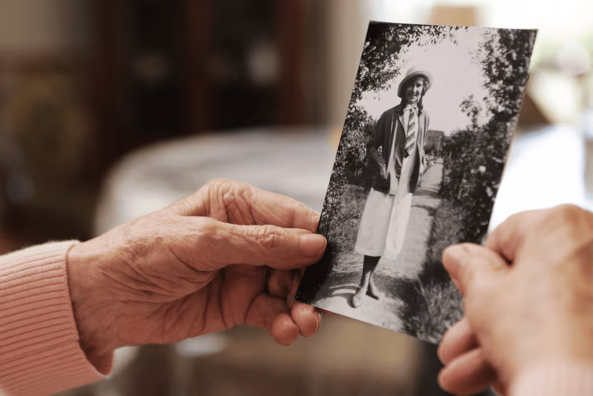 Close-up of elderly hands holding a black and white vintage photograph of a woman standing outdoors on a path, wearing a hat, jacket, and skirt.