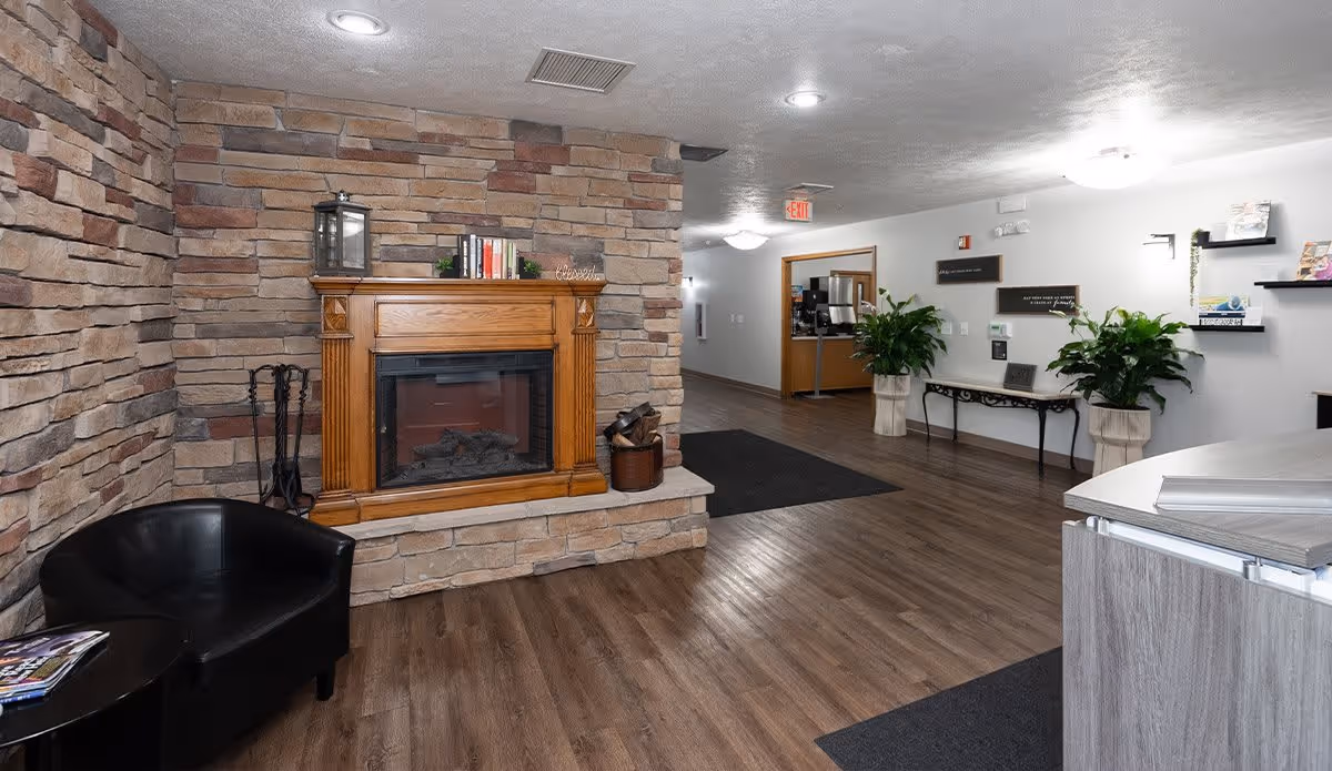 Interior view of a senior living facility lobby area featuring a stone wall with a wooden-framed fireplace, a black leather chair, a small round table with magazines, wooden flooring, potted plants, wall shelves with decorative items, and a hallway leading to other rooms.