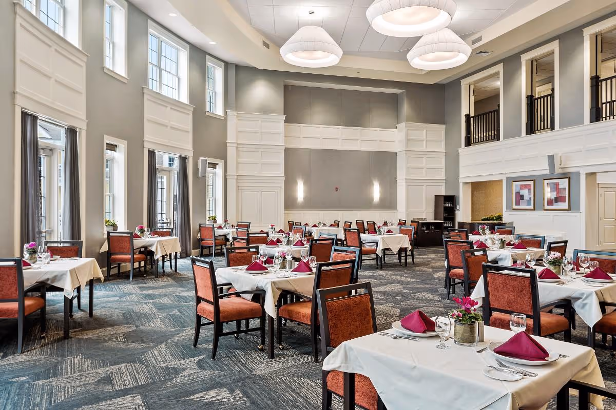 A spacious, elegant dining room with multiple tables covered in white tablecloths, each set with wine glasses, plates, silverware, and folded burgundy napkins. The room features high ceilings with large, modern light fixtures, tall windows with gray curtains, and a patterned carpet floor. There are decorative plants on the tables and framed artwork on the walls.
