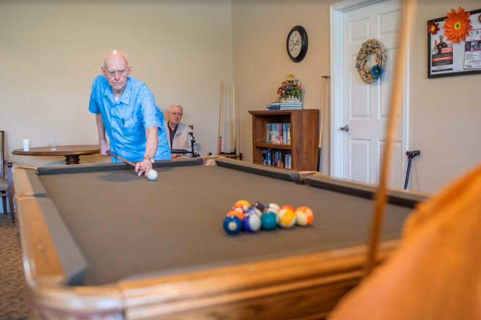 Two elderly men playing pool in a recreation room with a pool table, bookshelf, wall clock, and a door in the background.