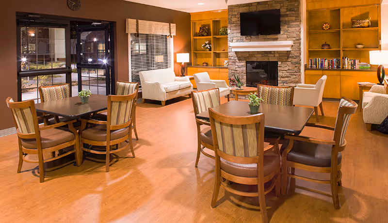 A cozy common area in Pisgah Valley Retirement Community featuring wooden tables with chairs, a stone fireplace with a mounted flat-screen TV above it, built-in wooden shelves with decorative items and books, and comfortable armchairs and sofas arranged around the room. Large windows with blinds and a glass door provide natural light.