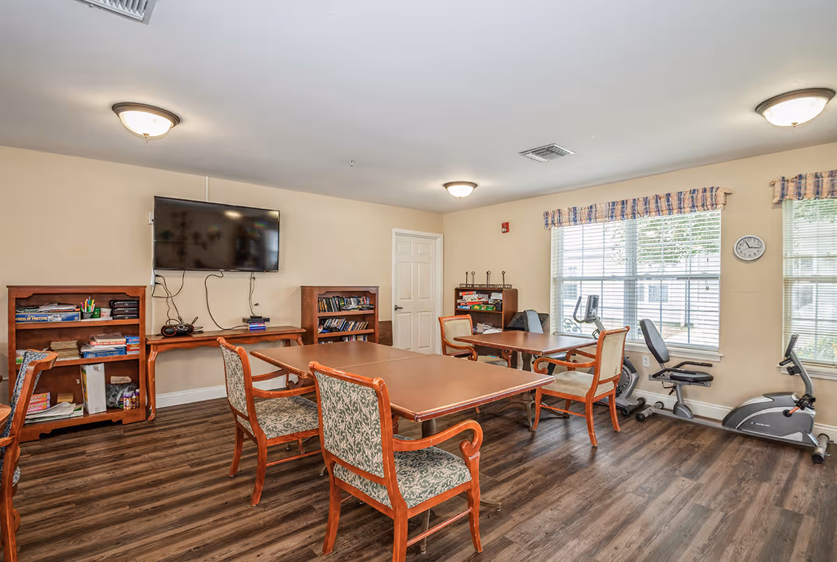 A well-lit activity room with wooden tables and cushioned chairs arranged in the center. The room features a wall-mounted flat-screen TV, bookshelves filled with board games and books, and two exercise bikes near windows with blinds and valances. The floor is wood laminate, and the walls are painted beige.