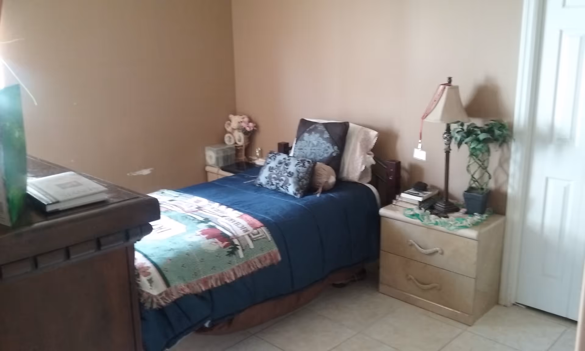 A small bedroom with a single bed covered in a blue bedspread and decorative pillows. There is a nightstand on each side of the bed; one has a lamp, books, and a small potted plant, while the other has a clock and a flower arrangement. The walls are beige, and the floor is tiled.