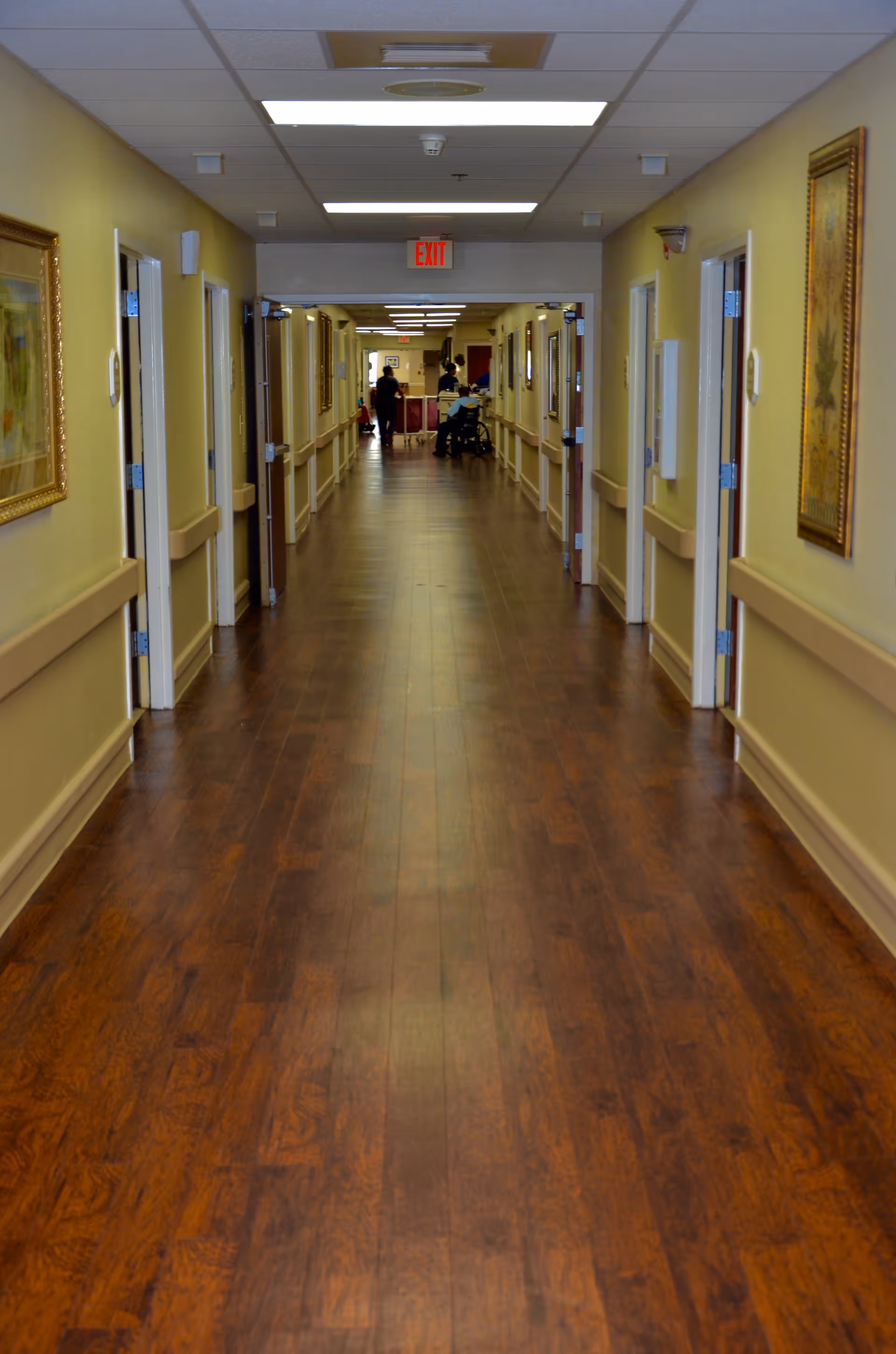 A long, well-lit hallway in a healthcare facility with wooden flooring and beige walls. Several open doors line both sides of the corridor, and framed artwork hangs on the walls. At the far end, a few people, including one in a wheelchair, are visible near a reception or nurse station. An exit sign is mounted above the doorway at the end of the hallway.