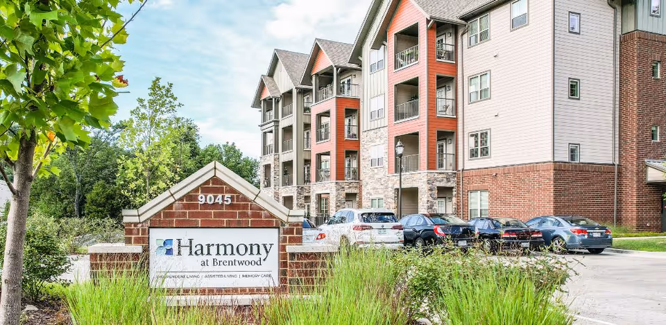 Exterior view of Harmony at Brentwood senior living facility showing a multi-story building with balconies, parked cars, and a brick sign with the facility's name and address 9045 surrounded by greenery.