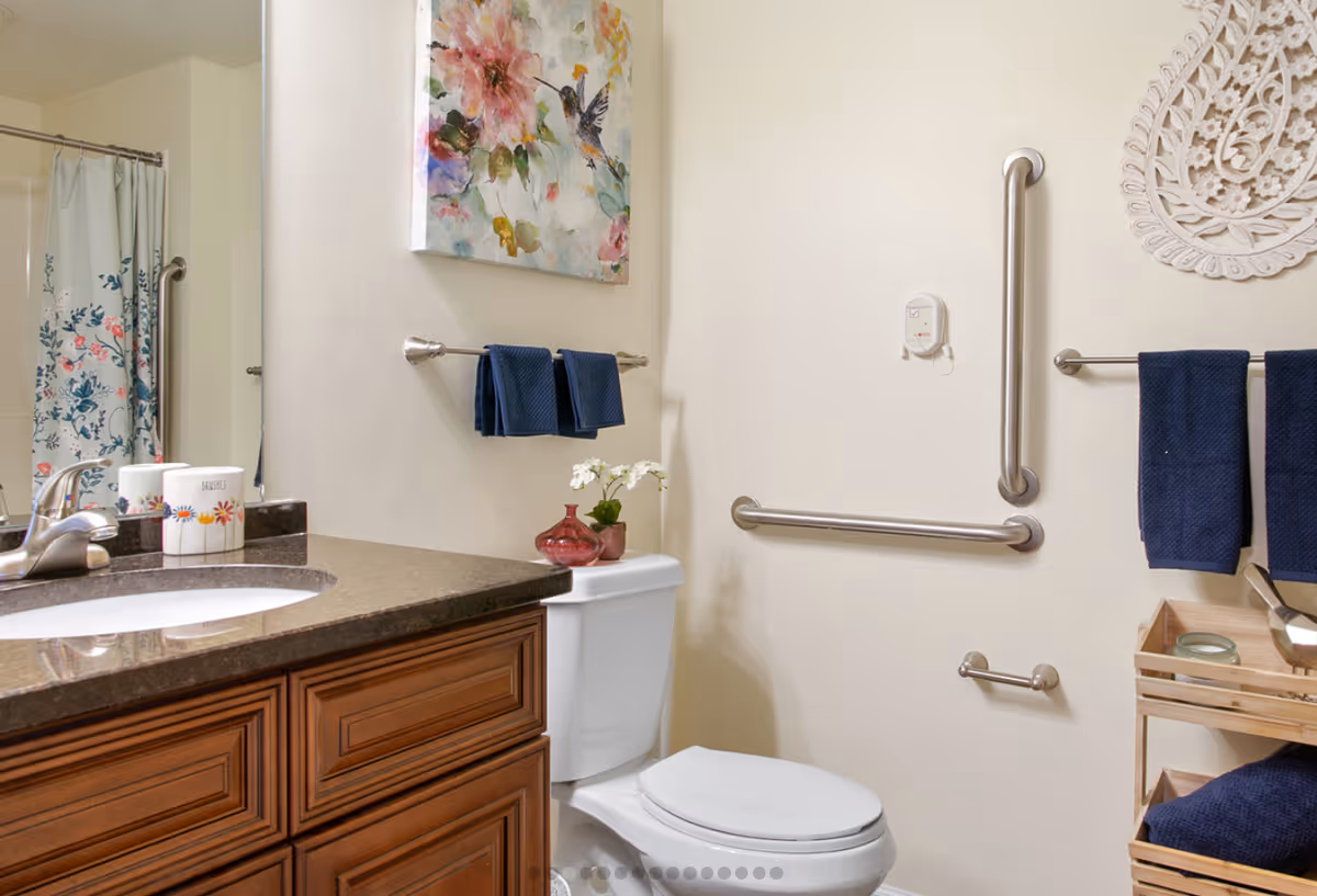 A bathroom with a wooden vanity and brown countertop featuring a sink and faucet. There is a white toilet with a small red vase and white flowers on top. The walls are light-colored with two towel racks holding dark blue towels. A floral shower curtain is visible in the mirror reflection. The bathroom has safety grab bars installed on the walls and decorative wall art above the toilet and towel rack.