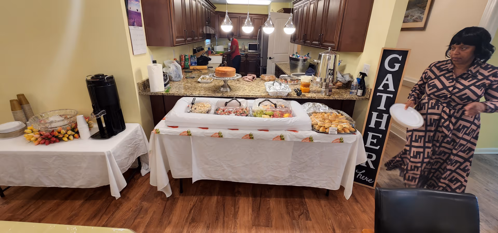 A dining setup in a kitchen area with tables covered in white tablecloths displaying various foods including a cake, fruit, sandwiches, and beverages. A woman holding a plate stands near a sign that reads 'GATHER here'. The kitchen has wooden cabinets and granite countertops.
