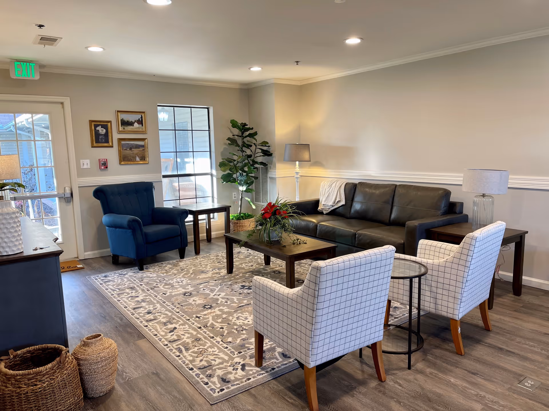 A cozy living room area with a dark leather sofa, two white checkered armchairs, a blue armchair, a wooden coffee table with a floral arrangement, a patterned area rug, two side tables with lamps, a large potted plant, and framed pictures on the wall near a window and an exit door.