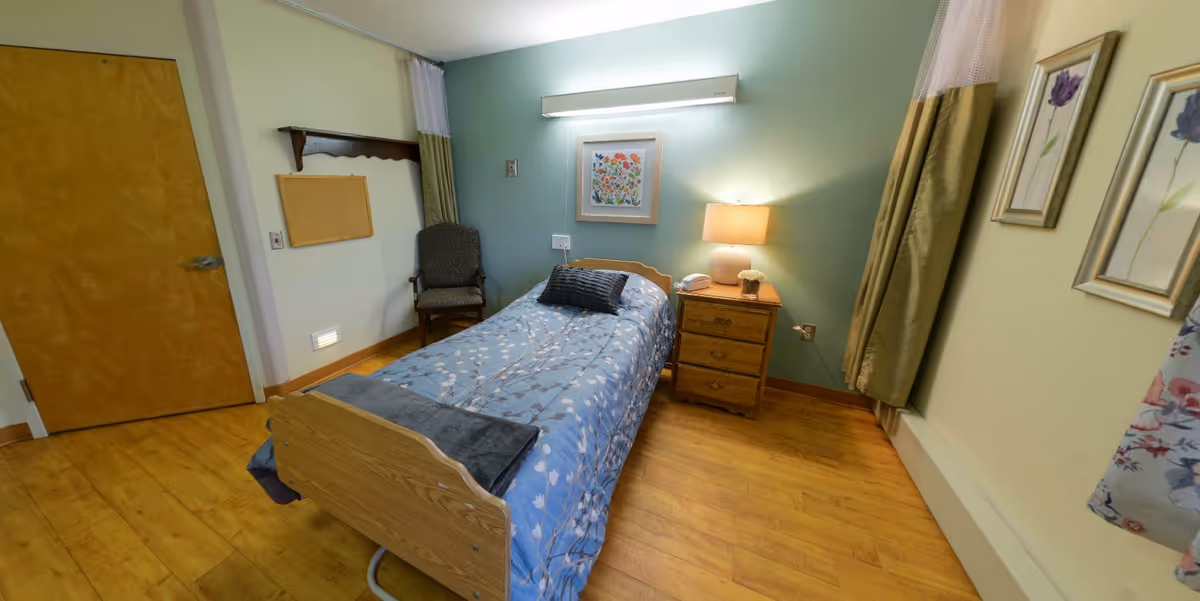 A small, cozy bedroom with a single bed covered in a blue floral bedspread, a wooden nightstand with a lamp and a telephone, a cushioned chair, and framed floral artwork on the walls. The room has light green and beige walls, wooden flooring, and a wooden door.