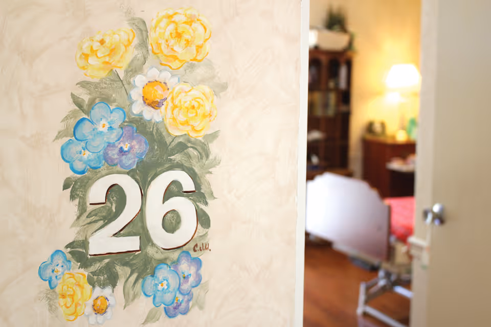 A wall painted with the number 26 surrounded by yellow and blue flowers. In the background, a partially open door reveals a cozy room with a bed, a wooden cabinet, and a lit table lamp.