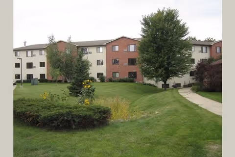 Exterior view of a three-story senior living facility building with a combination of brick and light-colored siding. The building is surrounded by well-maintained green lawns, bushes, trees, and a paved walkway leading to the entrance.