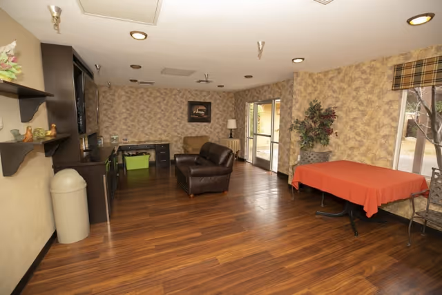 Interior room with wood flooring, patterned wallpaper, a brown leather armchair, a beige armchair, a table covered with a red tablecloth, metal chairs, a potted plant, and a large window with a view outside. Shelves with decorative items and a trash can are visible on the left side.