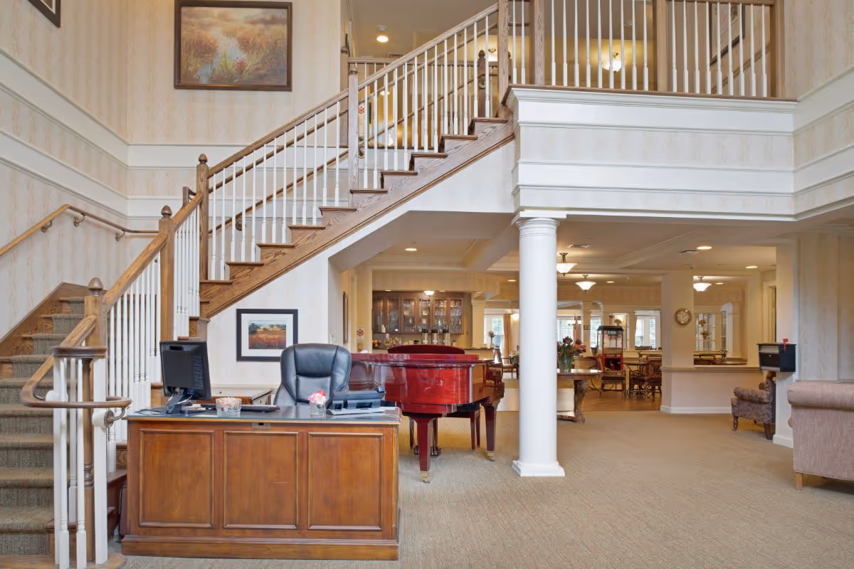 Interior view of a senior living facility lobby area with a wooden reception desk and black office chair in the foreground. Behind the desk is a staircase with wooden handrails and white balusters leading to an upper floor. A red grand piano is positioned under the staircase. The background shows a spacious common area with tables, chairs, and decorative lighting fixtures. The walls are decorated with framed paintings and light-colored wallpaper.
