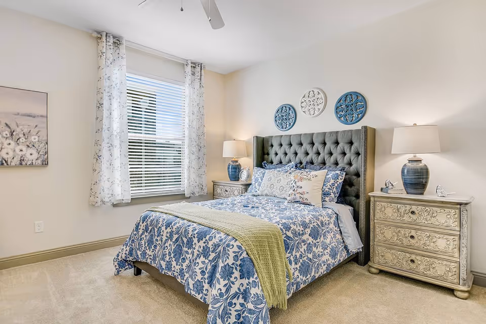 A cozy bedroom with a tufted dark gray headboard bed dressed in blue and white floral bedding and a light green throw blanket. There are two ornate cream-colored nightstands on either side of the bed, each with a blue ceramic lamp and decorative items. A window with white blinds and floral curtains is on the left wall, and three decorative circular wall hangings in blue and white are above the bed. The room has beige carpet and light-colored walls.
