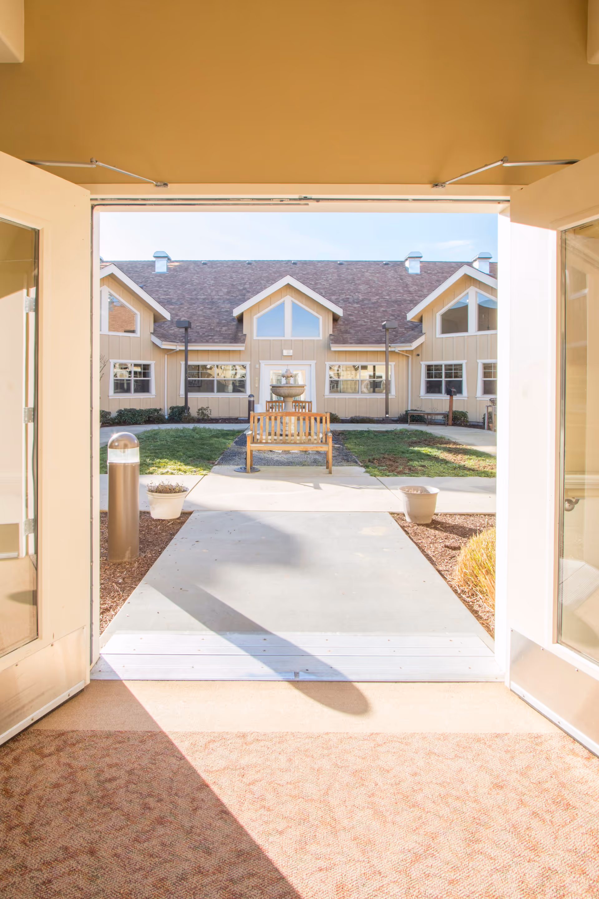 View through open double doors onto a courtyard with a bench and fountain in front of a light-colored memory care building.