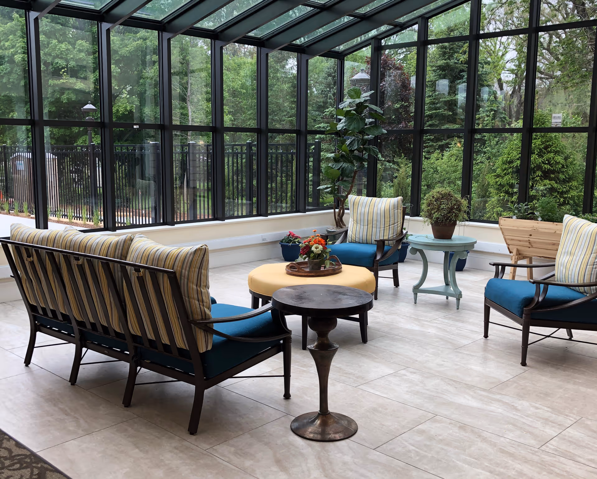 A bright sunroom with large floor-to-ceiling windows showing green trees outside. The room contains a seating area with a striped cushioned sofa, two armchairs with blue cushions and striped pillows, a round yellow ottoman with a tray of flowers, a small round wooden table, and two side tables with potted plants.