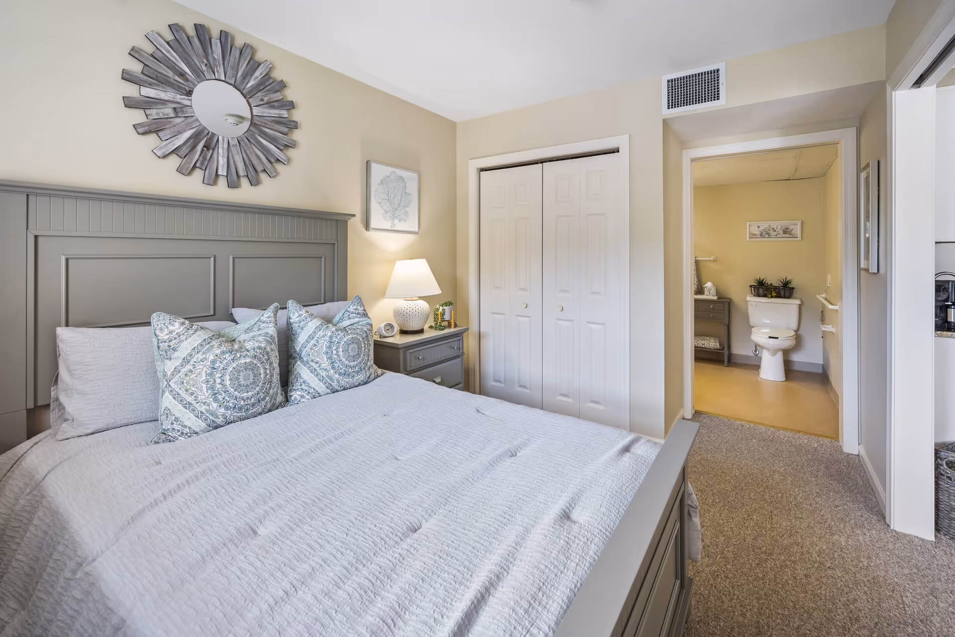 A bedroom with a gray bed frame and headboard, a neatly made bed with patterned pillows and a light gray bedspread. A nightstand with a lamp, clock, and small plant is next to the bed. There is a decorative round mirror on the wall above the headboard and a framed picture on the adjacent wall. A closet with white double doors is visible, and an open doorway leads to a bathroom with a toilet and small cabinet with plants.
