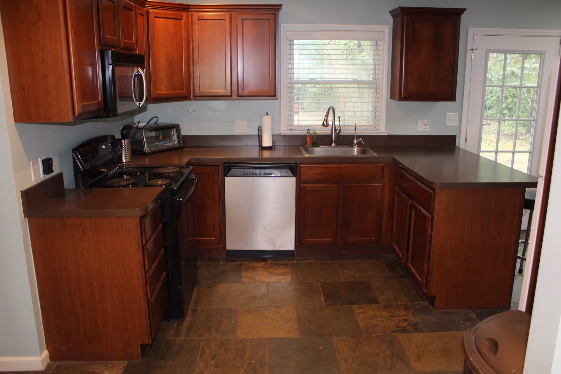 A kitchen with dark wood cabinets, a black electric stove, a stainless steel dishwasher, a double sink under a window with white blinds, a toaster oven on the counter, and a door leading outside. The floor is tiled with large stone tiles in various shades of brown.