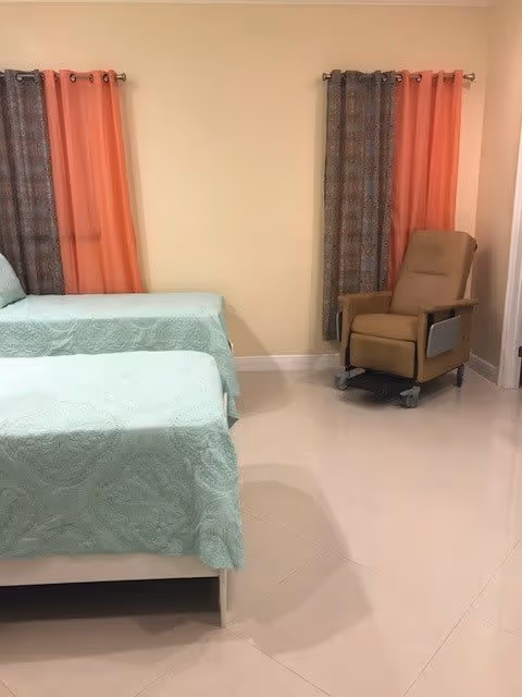 A room with two single beds covered in light green bedspreads, two windows with orange and patterned brown curtains, and a beige recliner chair on wheels positioned near the corner.