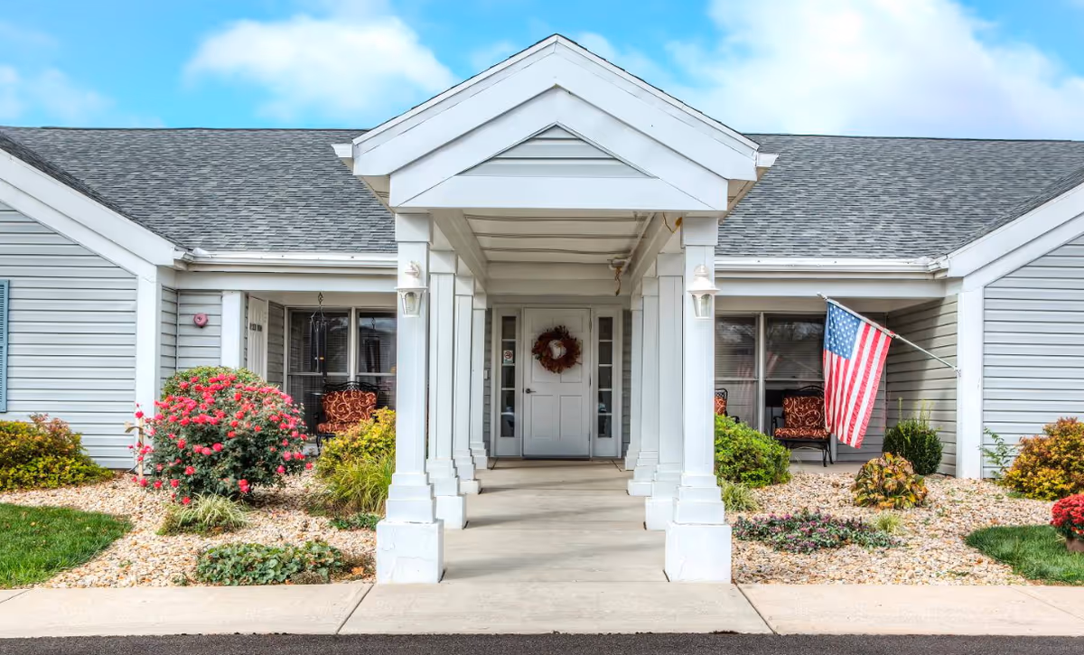 Front entrance of a single-story building with a covered walkway supported by white columns. The building has gray siding, a shingled roof, and a white door decorated with a wreath. There are landscaped flower beds with bushes and flowers on either side of the walkway, and an American flag is displayed on the right side near a window with two chairs.