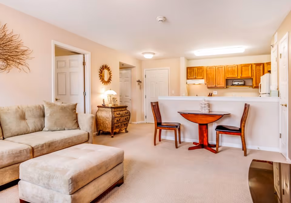 A cozy living room area with a beige sofa and matching ottoman, a small wooden table with two chairs, and a kitchen with wooden cabinets in the background. The room has beige walls and carpet, a decorative mirror, and a small chest with a lamp on it.