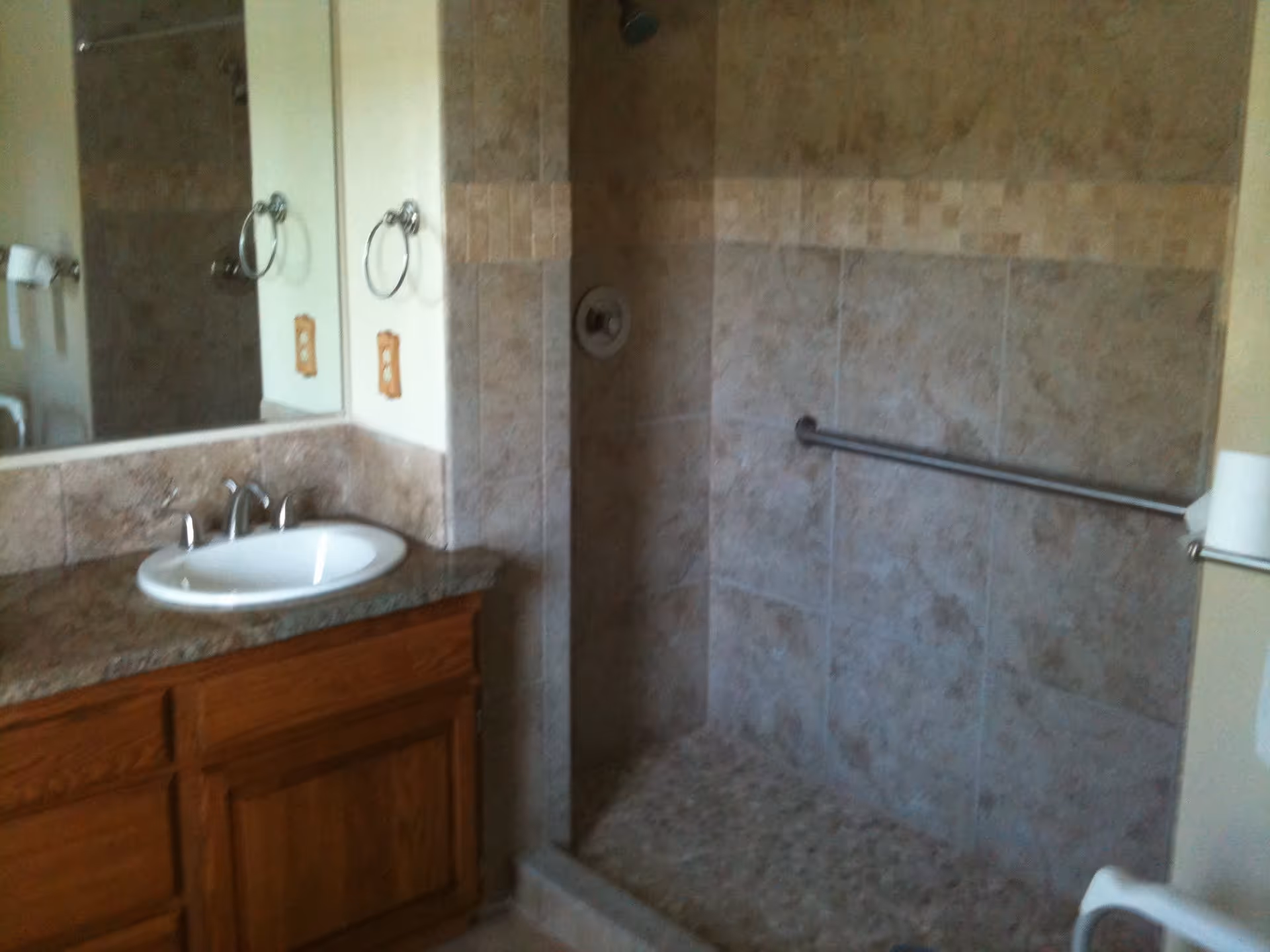 Bathroom with a wooden vanity featuring a white sink and granite countertop, a large mirror above the sink, and a tiled walk-in shower with a grab bar on the wall.