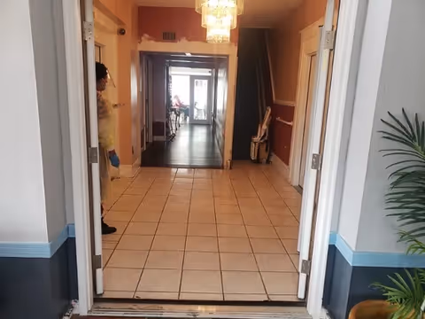 Indoor hallway of a senior care facility with tiled floor, doors, and people at far end.