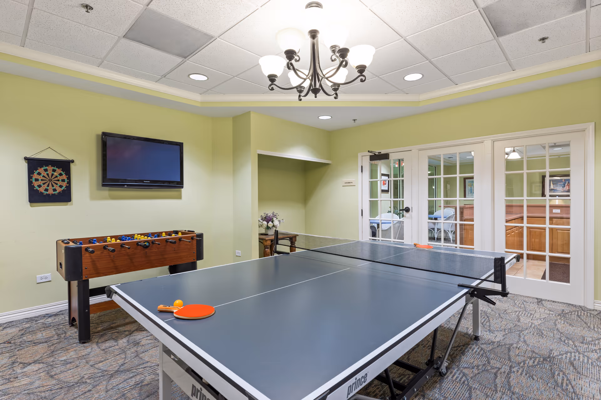 Indoor recreational room with a ping pong table in the center, a foosball table against the wall, a dartboard, and a wall-mounted TV. The room has light green walls, a chandelier, and glass double doors leading to another room with seating.