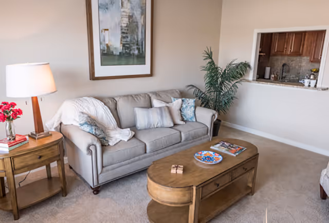 Bright living room with a beige sofa, wooden coffee and side tables, a lamp, artwork on the wall, and a pass-through window to the kitchen.