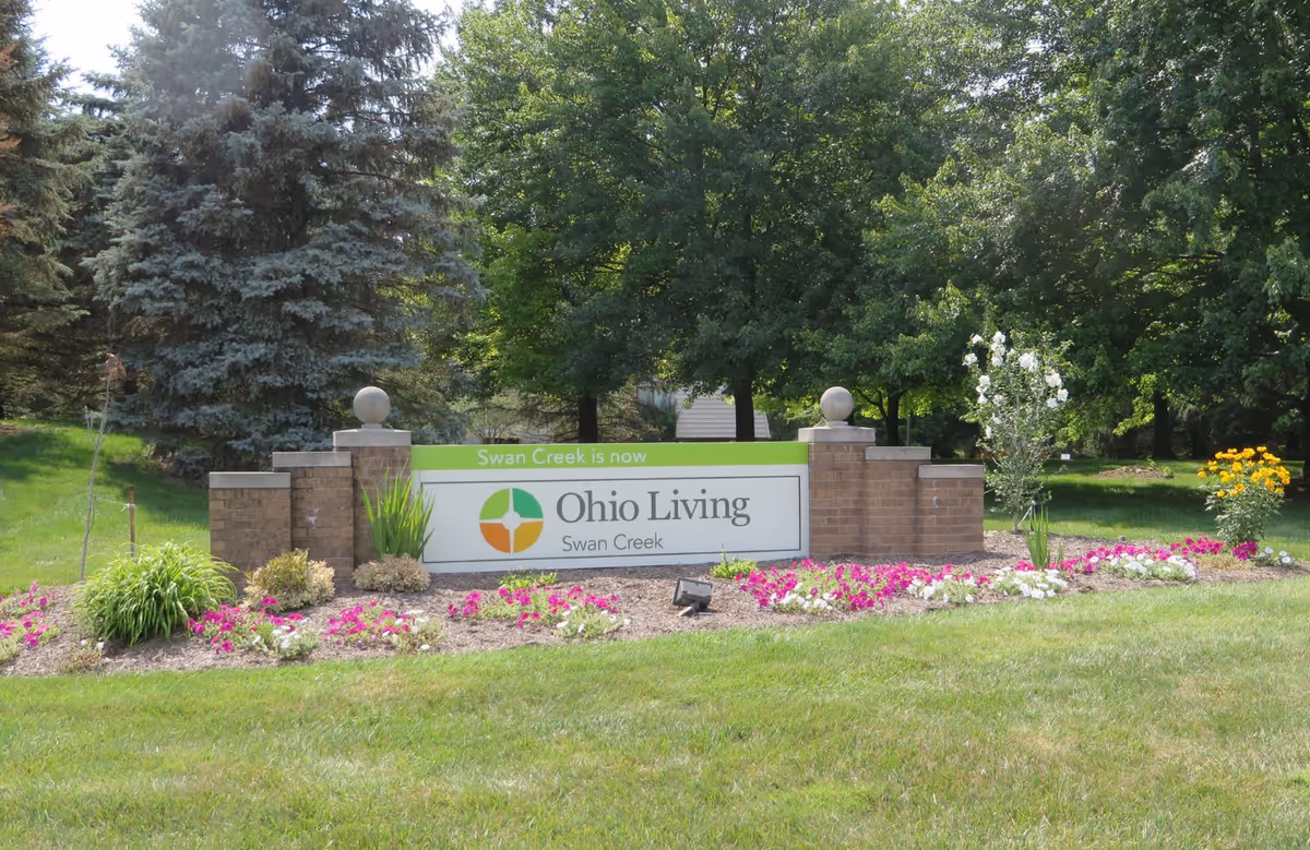 Outdoor view of a landscaped entrance sign for Ohio Living Swan Creek, surrounded by green grass, colorful flowers, and trees in the background.