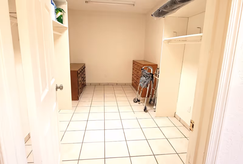 A bright room with white tiled floor and cream-colored walls, featuring two wooden dressers against the far wall and a walker with a patterned bag hanging on it. There is an open closet space on the right side with a hanging rod and shelf, and a partially open door on the left side.