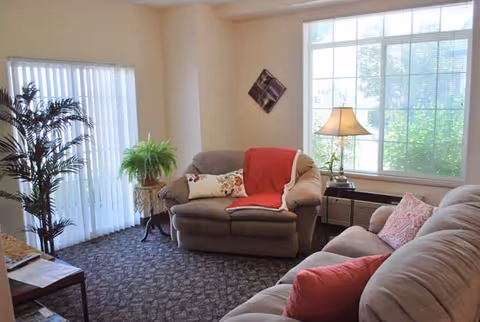 A cozy living room with two beige sofas adorned with red and floral pillows and a red throw blanket. There is a large window letting in natural light, a table lamp on a side table, a potted plant near sliding glass doors with vertical blinds, and a small decorative wall piece.