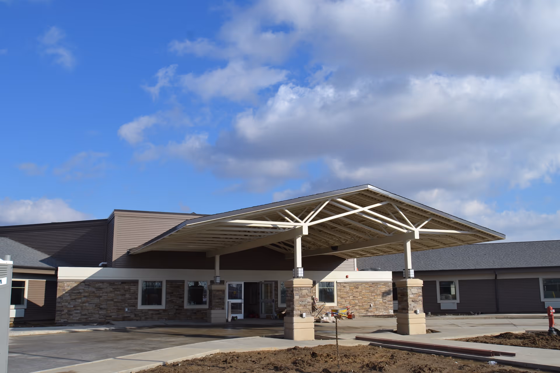 Exterior view of a single-story building with a large covered entrance supported by stone and beige pillars under a partly cloudy blue sky.