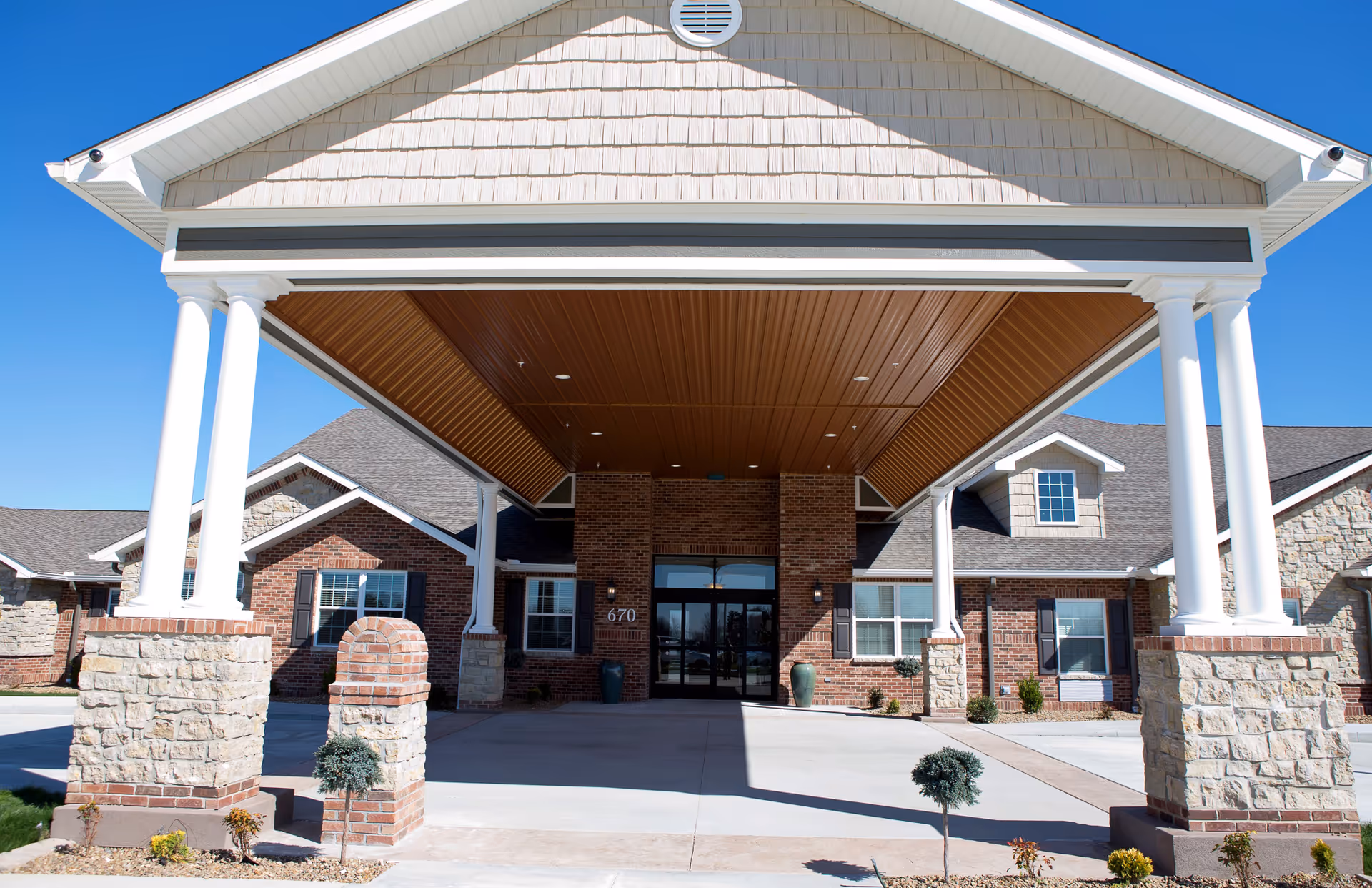 Front entrance of The Villas of Jackson facility showing a covered driveway with white columns and stone bases, brick walls, windows with shutters, and a clear blue sky.