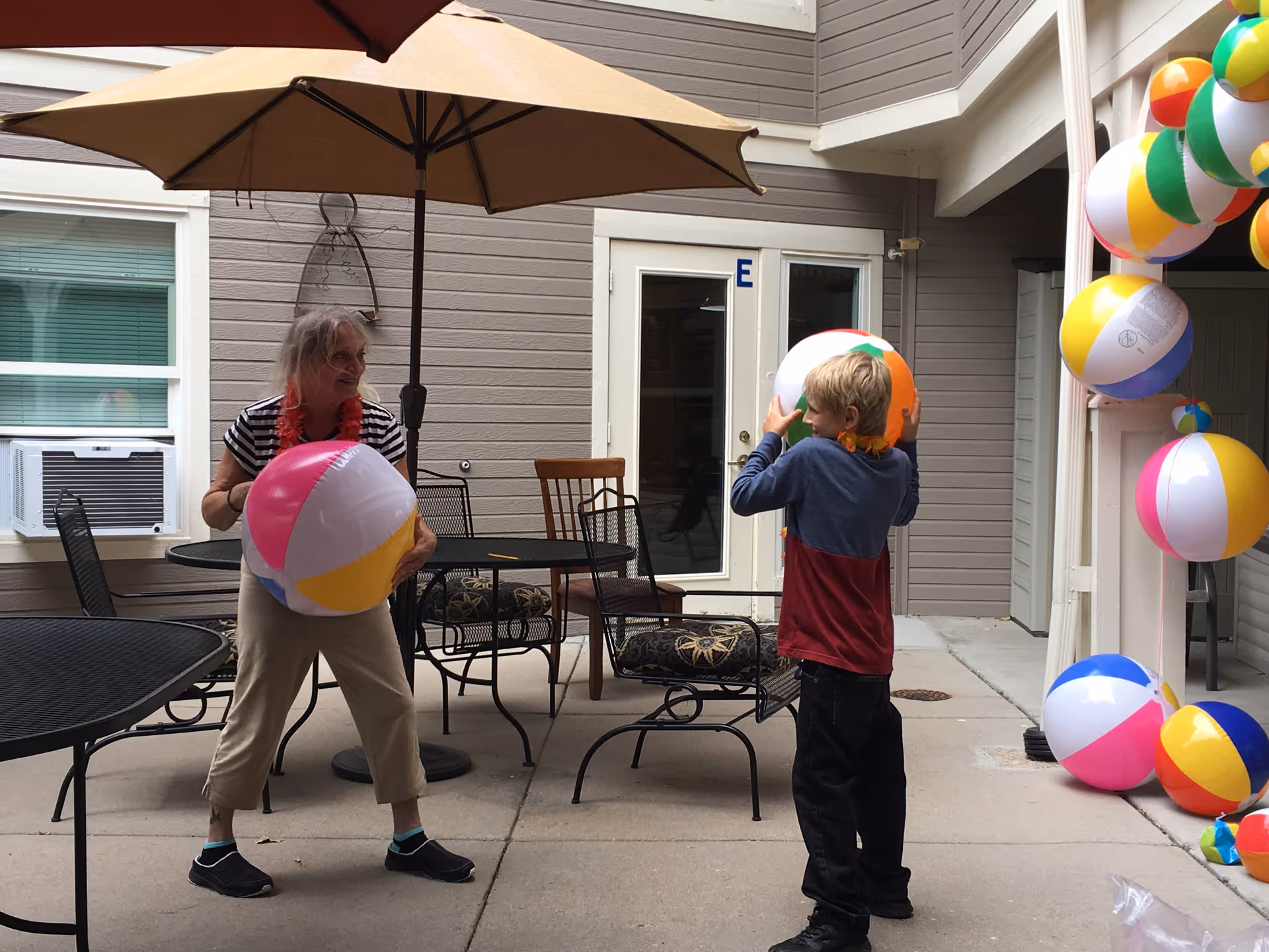 An elderly woman and a young boy play with colorful beach balls in an outdoor courtyard area of a senior living facility. The courtyard has patio tables, chairs, and large umbrellas. The building exterior is visible with windows and a door marked with the letter E. Several beach balls are hanging on the right side of the image.